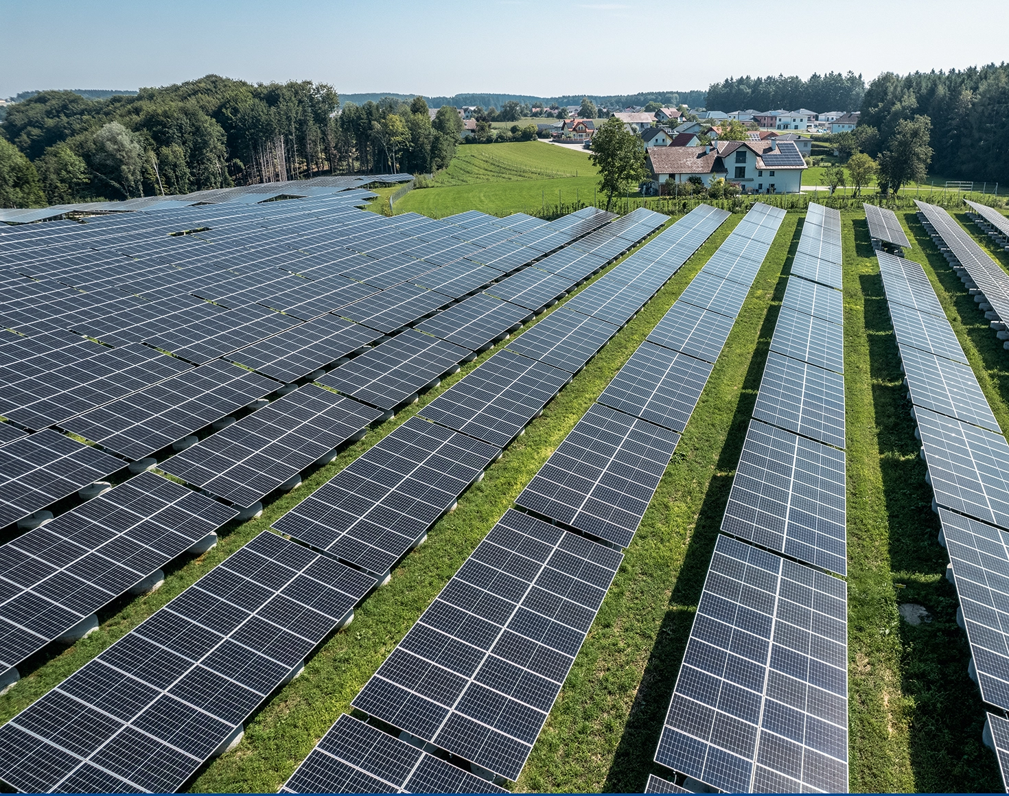 A view from above of a large-scale photovoltaic system. The solar park is surrounded by lush greenery. Small picturesque villages can be seen in the background.