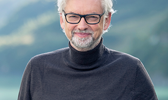 VERBUND CEO Michael Strugl smiles into the camera and stands in front of a blurred mountain backdrop in the sunshine wearing a black turtleneck sweater